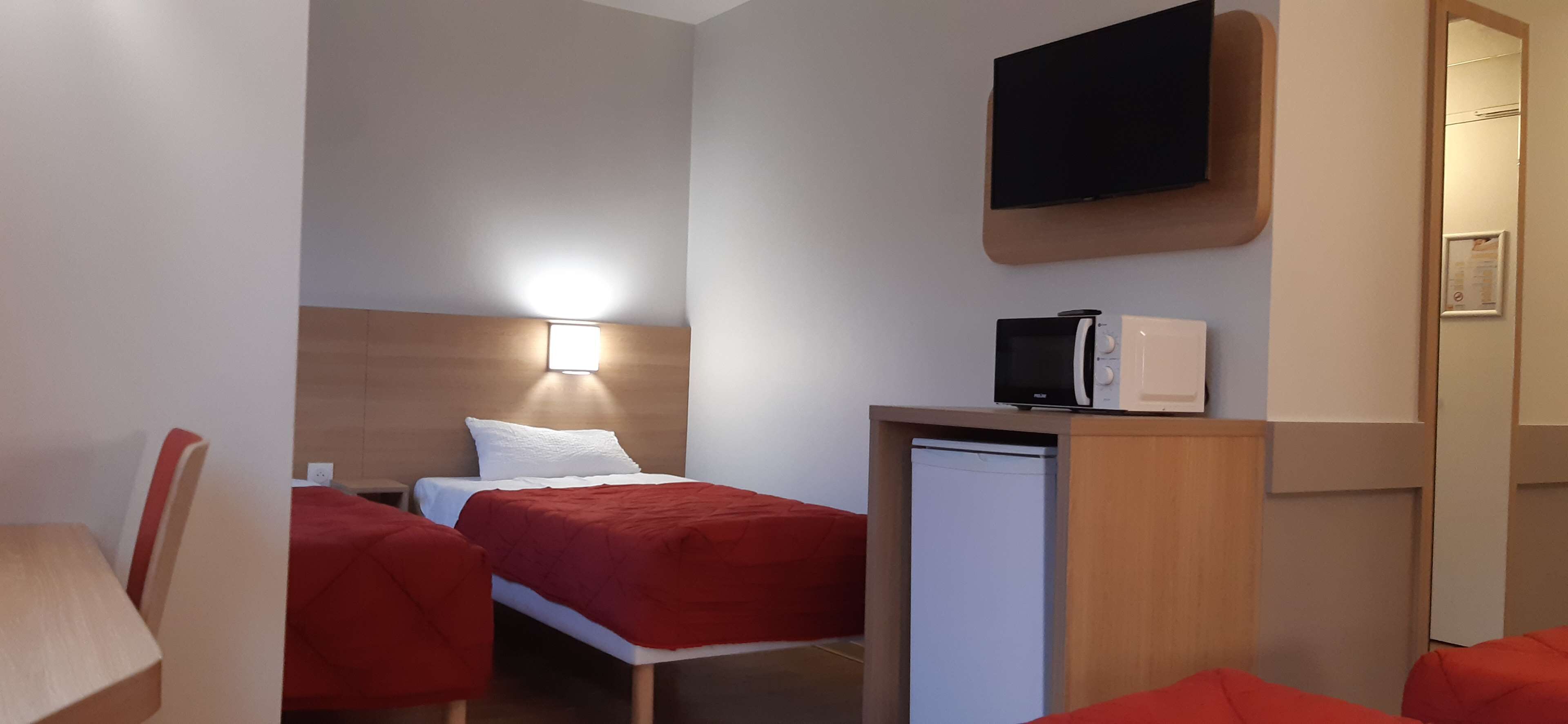 A compact hotel room with a bed, a desk with a microwave, a TV mounted above the desk, and a small refrigerator.