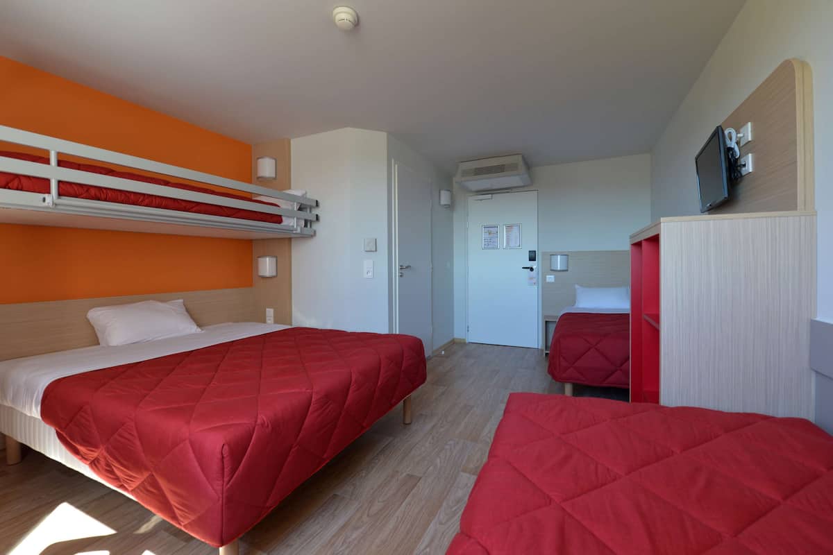Ein Hotelzimmer mit zwei Betten, eines mit roter Bettdecke und das andere mit weißer Bettdecke. Es gibt eine weiße Tür mit kleinem Fenster, eine an der Wand montierte Klimaanlage und ein an der Wand über der Tür angebrachtes Fernsehgerät.