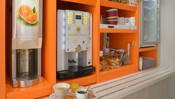 Estación de desayuno con cafetera, dispensador de jugo de naranja y una variedad de alimentos, como pasteles y pan.