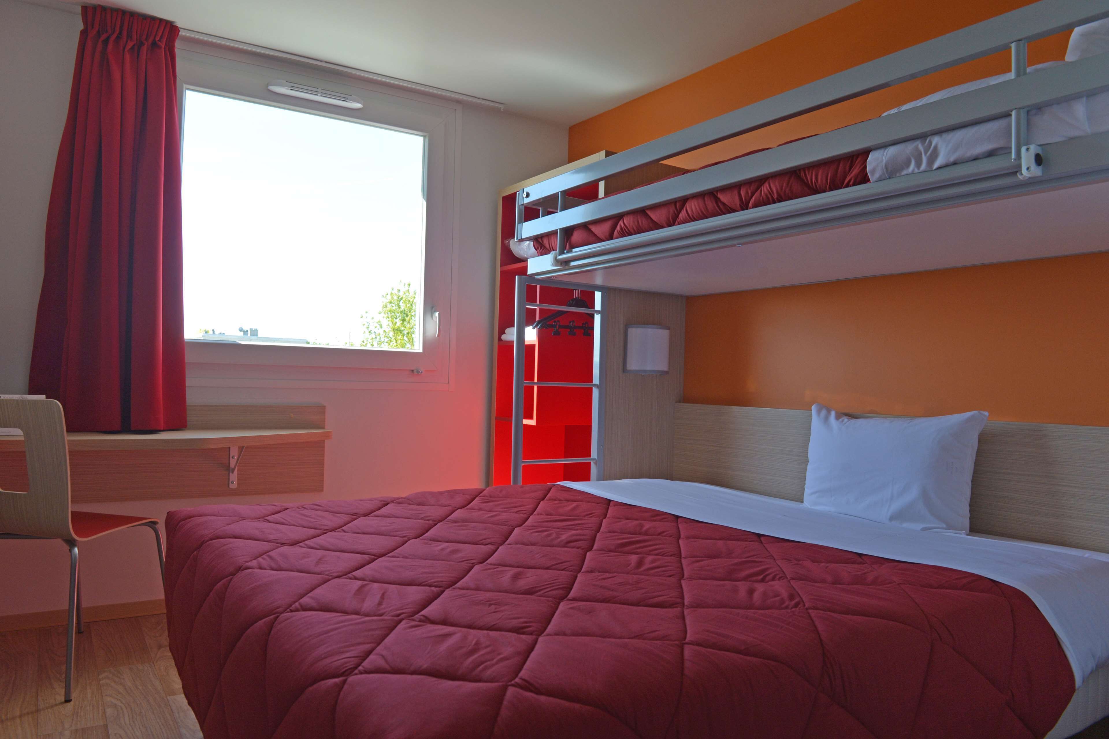 A bunk bed room with a red bedspread, a desk with a chair, and a window with red curtains.