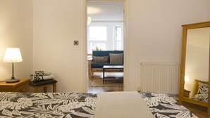 A bedroom with a bed, a nightstand, a lamp, a mirror, and a view into another room with a sofa and a window. - Sleep & Stay Oxford - Beautiful Flat Close to town (Oxford)