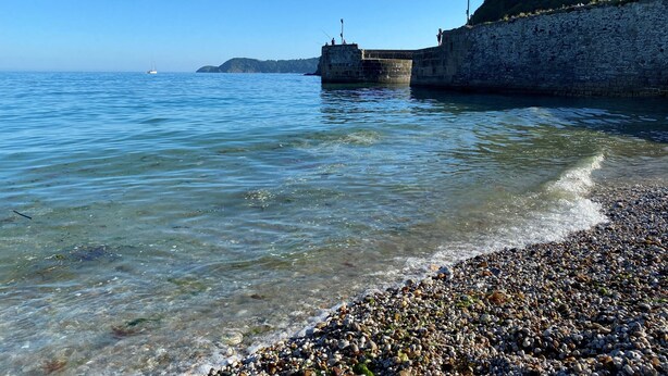 A clear coastal view with a pebbled beach, a stone wall, and a lighthouse in the distance.