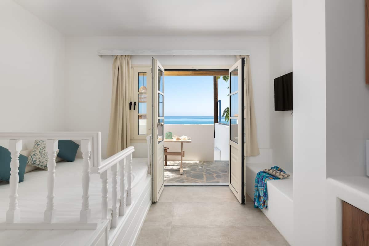 A bedroom with a view of the sea, a balcony with a table and chairs, and a TV mounted on the wall.