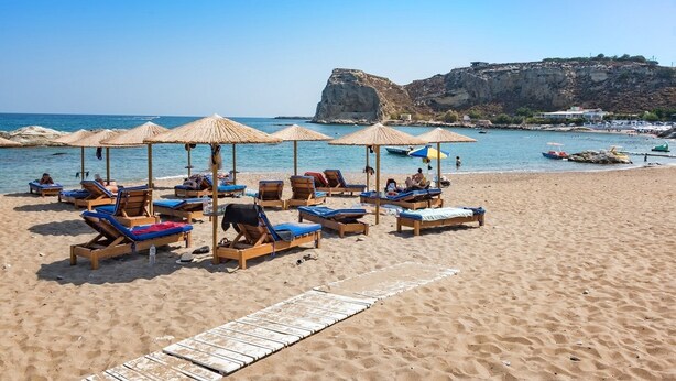 A beach with sun loungers and umbrellas, a clear blue sky, and a rocky cliff in the background. - Stegna Akti (Rhodes)