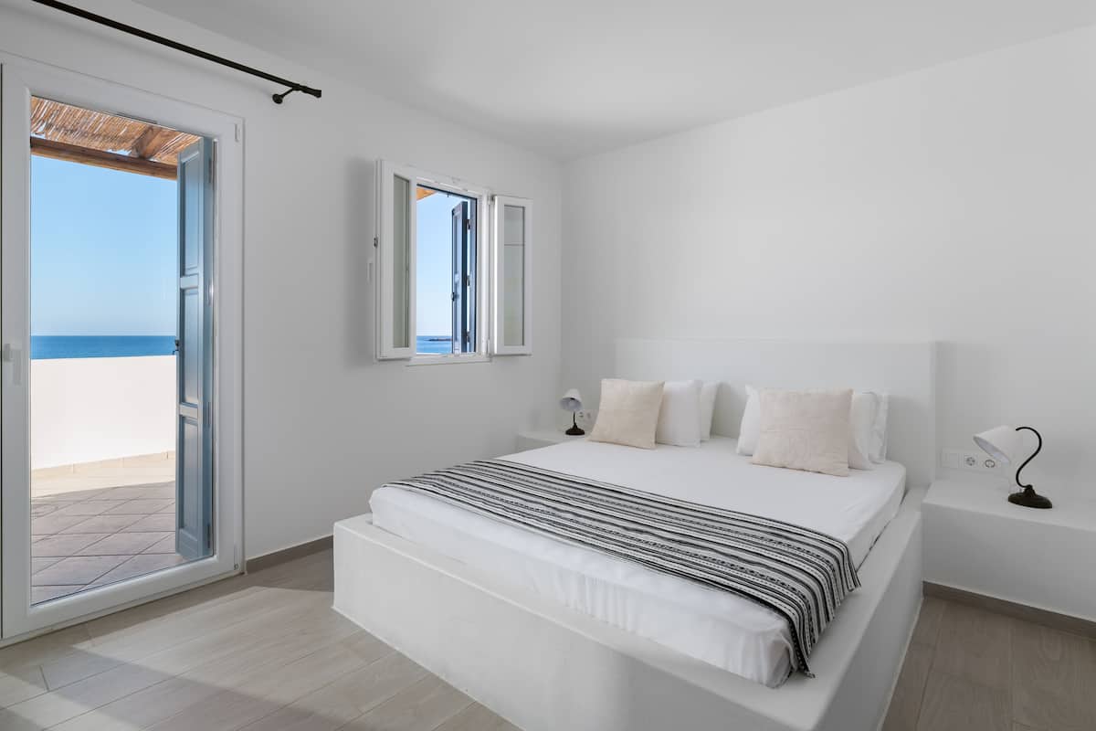A bedroom with a bed, a nightstand, a lamp, and a view of the beach through an open door.