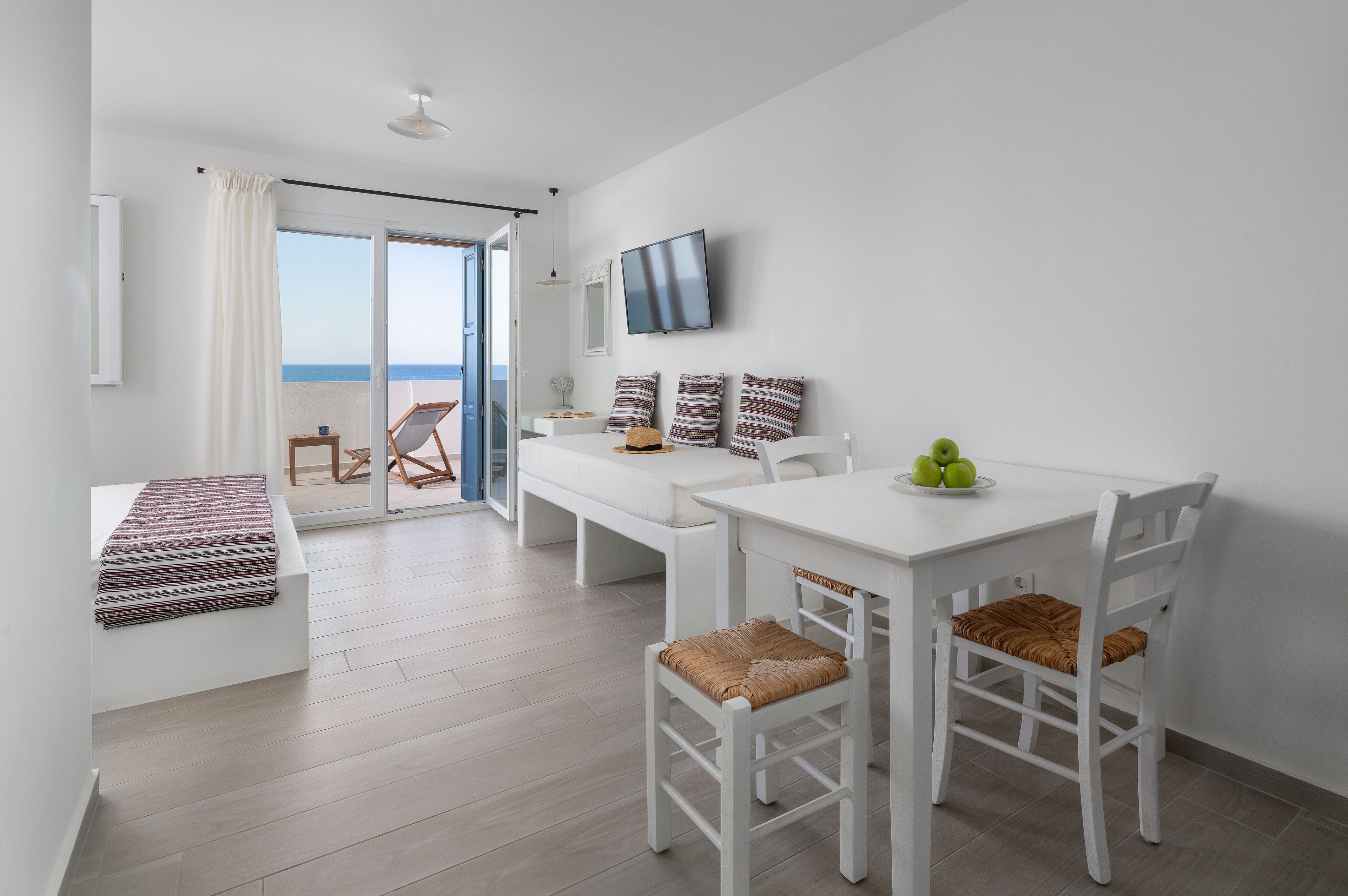 A modern, well-lit room with a white sofa, a dining table with chairs, a TV mounted on the wall, and a view of the sea through a glass door.