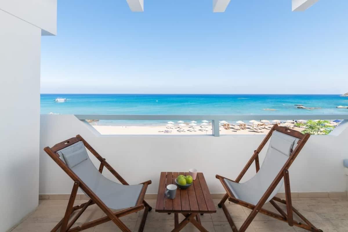 A balcony with two wooden chairs and a small table, overlooking a beach with clear blue water and a boat in the distance.