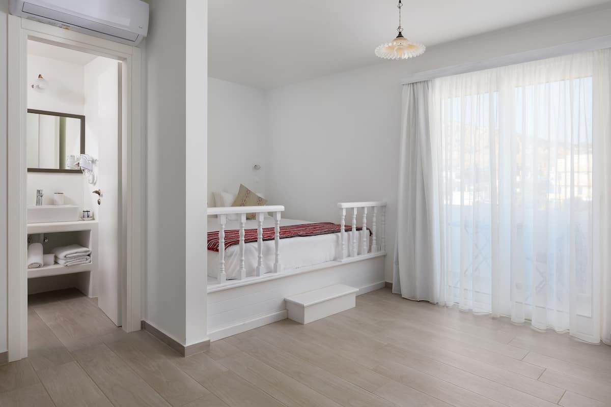 A bedroom with a white bed, a small bedside table, a window with sheer curtains, and a white wardrobe.
