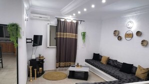 A modern living room with a sofa, coffee table, and a TV. There is a window with curtains, a plant, and wall decorations. - Relax in the comfort of Dar Elyas: our villa with swimming pool in Djerba! (Mezraya)