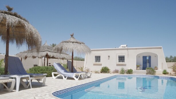 A pool area with lounge chairs and thatched umbrellas, a white building with a blue door, and a clear blue sky. - Relax in the comfort of Dar Elyas: our villa with swimming pool in Djerba! (Mezraya)