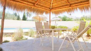 A covered outdoor seating area with a thatched roof, white metal chairs, and a table. - Relax in the comfort of Dar Elyas: our villa with swimming pool in Djerba! (Mezraya)