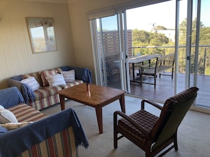Relax in this inviting living room with a sofa, armchair, and coffee table, leading to an outdoor deck with a table and chairs.