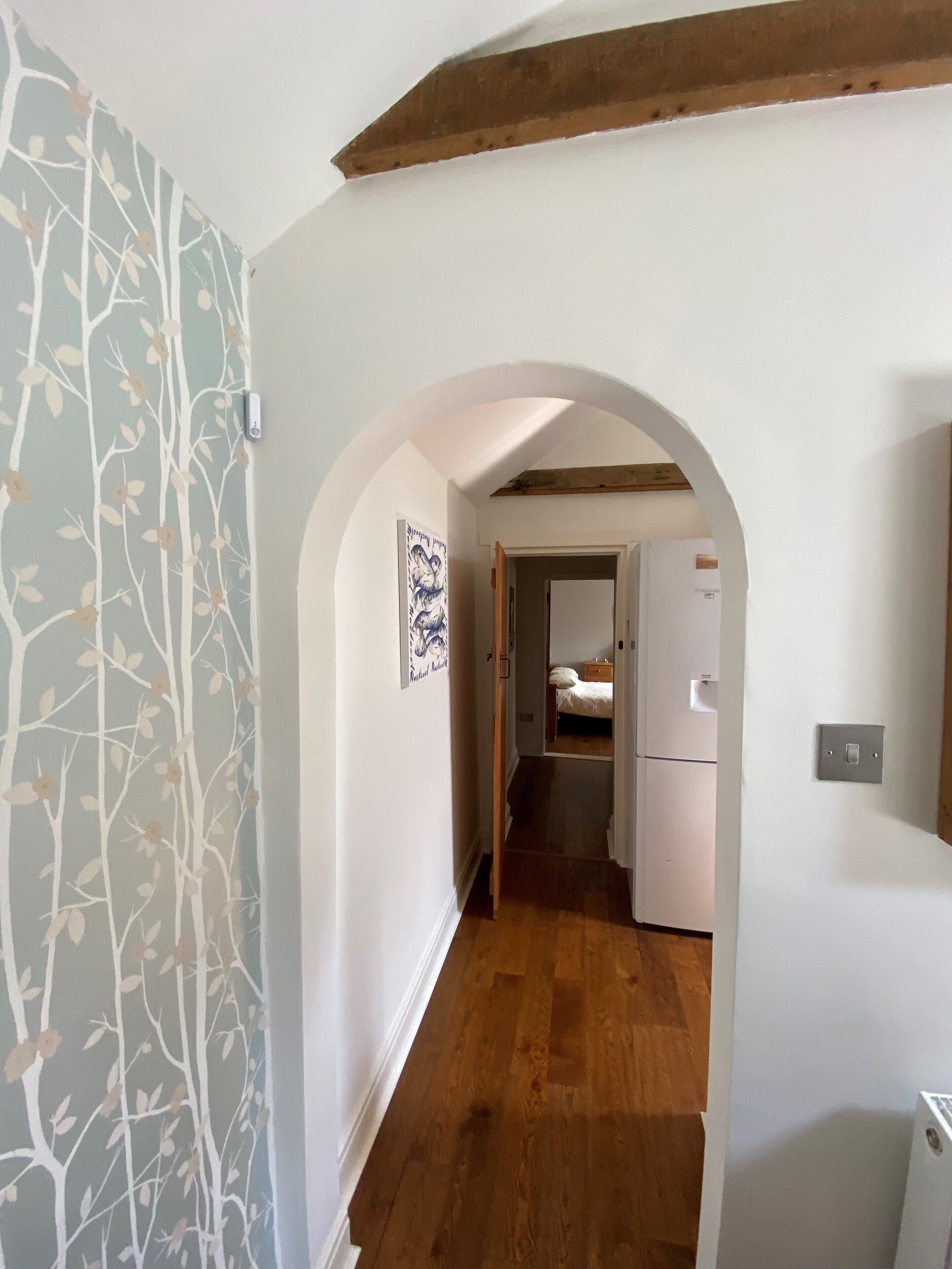 Un pasillo estrecho con suelo de madera, paredes blancas y un diseño de árboles decorativo en una pared. Un arco de entrada da a un dormitorio con una cama y un armario blanco.