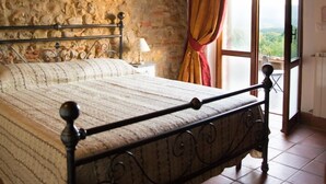 Ein Schlafzimmer mit Steinwand, einem Bett mit Metallgestell, einem Fenster mit Vorhängen und Blick ins Grüne.