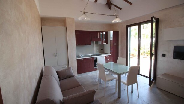 A modern living room with a sofa, a dining table with chairs, a kitchen area, and a TV mounted on the wall.