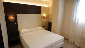 A neatly made bed in a hotel room with a headboard, two wall-mounted lamps, and a window with curtains.