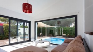 Una sala de estar moderna con un gran ventanal que da a una zona de piscina, una mesa de centro blanca y una lámpara colgante roja.