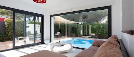 Una sala de estar moderna con un gran ventanal que da a una zona de piscina, una mesa de centro blanca y una lámpara colgante roja.