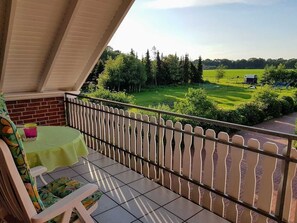 Uma varanda com mesa e cadeiras, vista para um campo verde e um veículo estacionado ao longe.