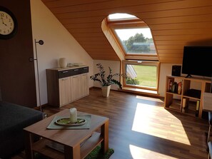Una sala de estar moderna con techo inclinado, suelo de madera, una mesa de comedor con un centro de velas, un sofá, un televisor sobre un mueble de madera y una estantería.