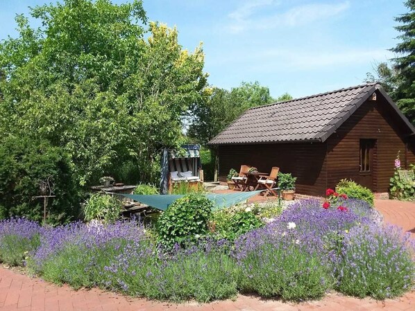 A wooden cabin with a tiled roof, surrounded by a garden with purple flowers and a small pool area. - Apartment - Apartment Hahn, 95129 (Rhauderfehn)