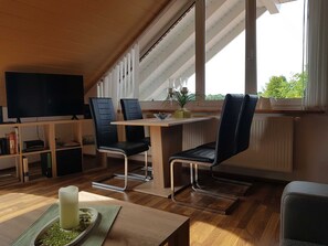 Uma sala de estar moderna com teto de madeira, uma TV de tela plana, uma mesa de jantar com cadeiras e uma janela grande com vista para o verde.