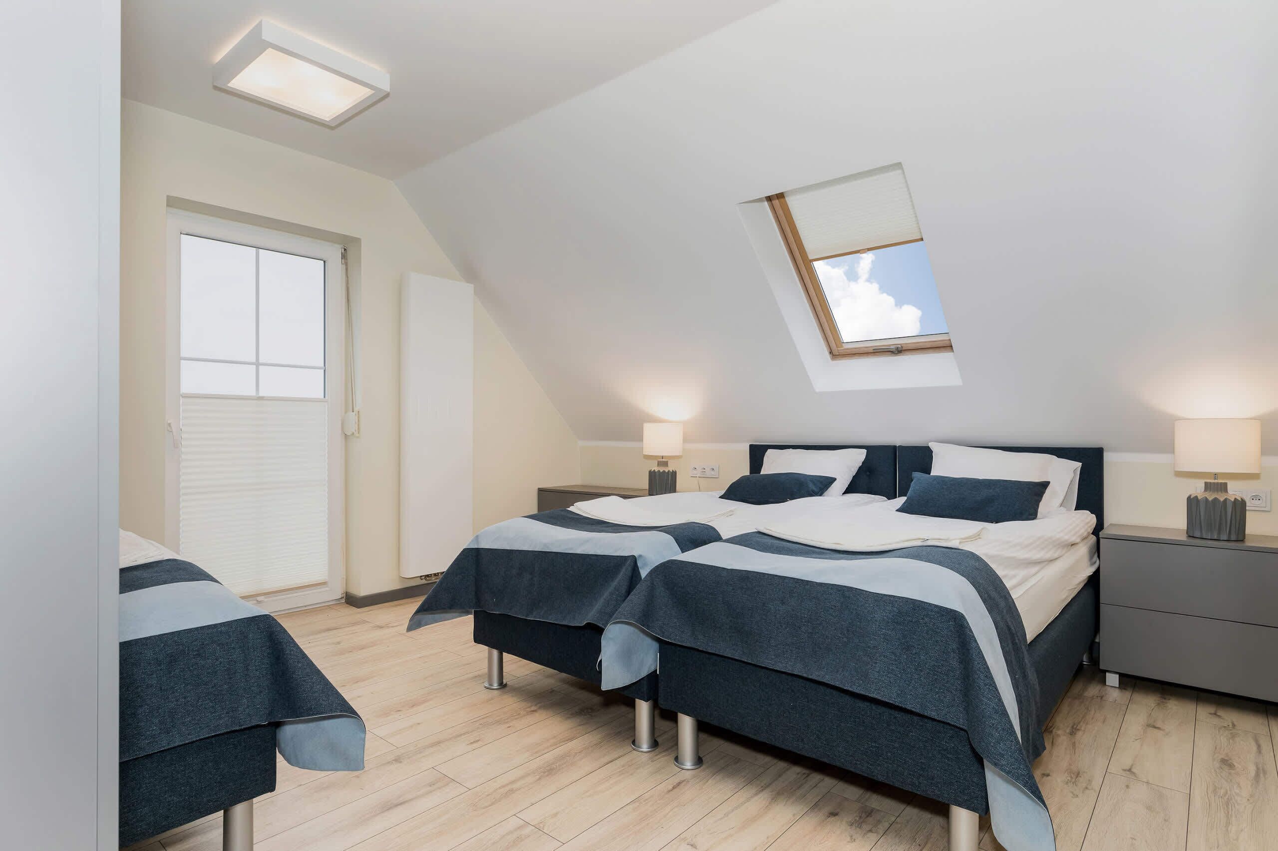 Ein Schlafzimmer mit schrägen Wänden, zwei Betten mit dunkler Bettwäsche, Nachttischlampen, einem Fenster mit Jalousien und einem kleinen Dachfenster.