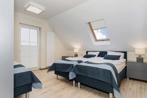 Un dormitorio con techo inclinado, dos camas con ropa de cama oscura, lámparas de noche, una ventana con persianas y un tragaluz pequeño.