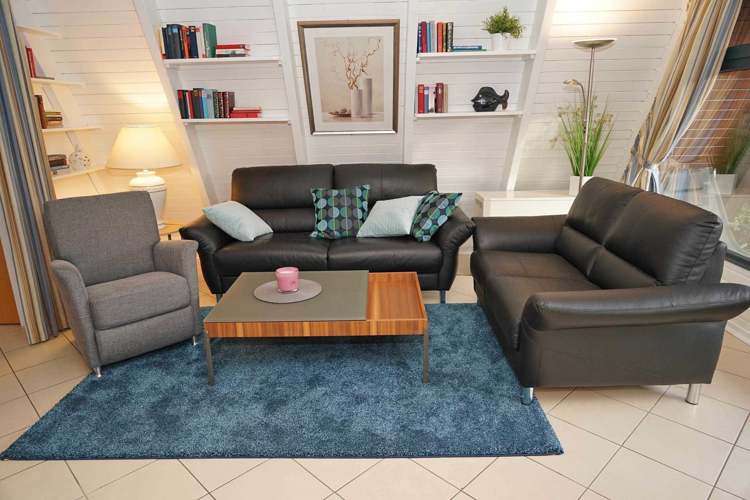 A modern living room with a black leather sofa, a grey armchair, and a wooden coffee table. There are bookshelves with books and decorative items, a framed artwork on the wall, and a lamp on a side table.