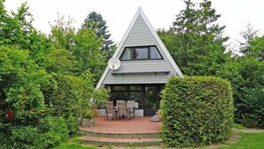 Een modern huis met een puntdak, omringd door weelderige begroeiing en een terras met tuinmeubilair.