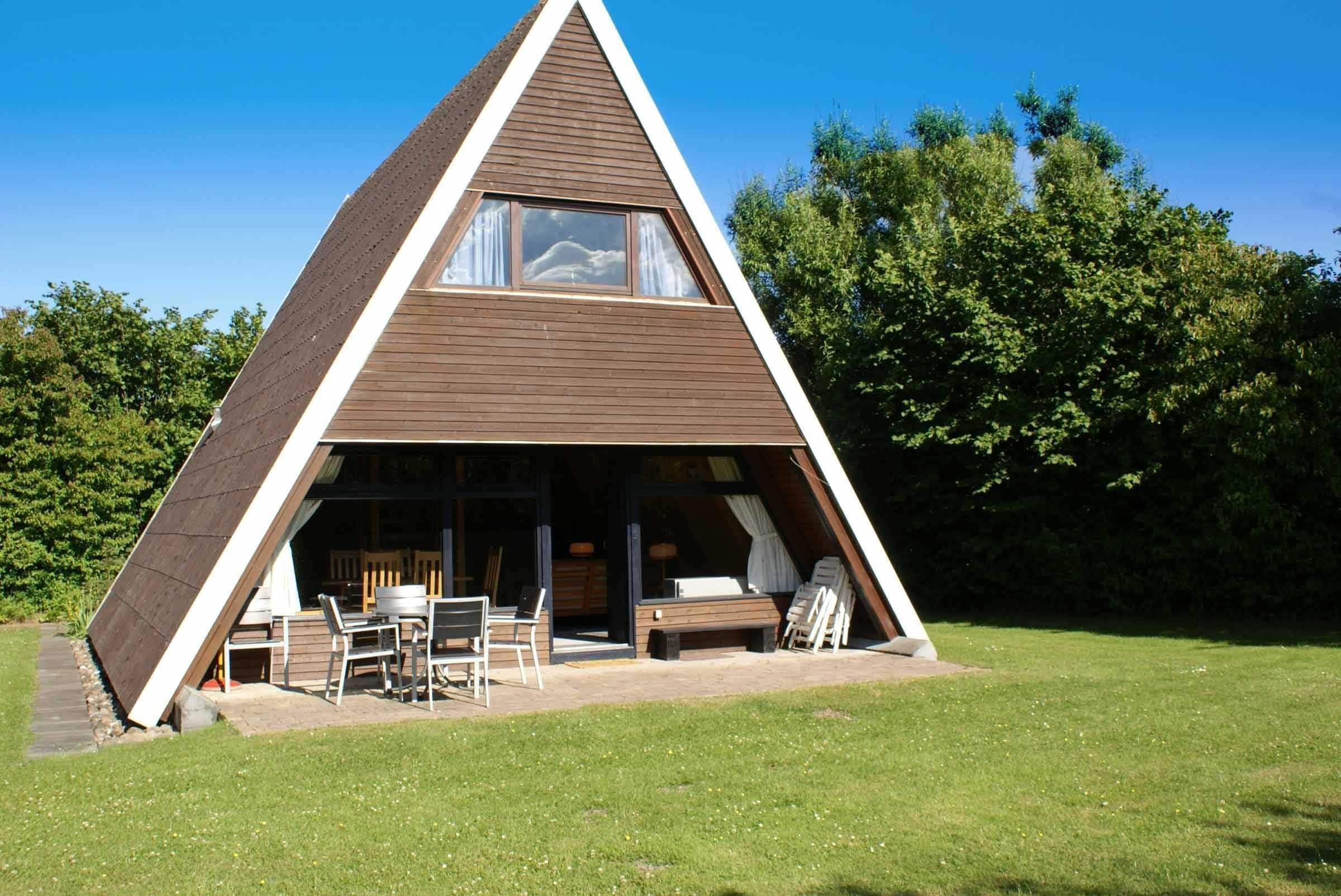 Uma casa de madeira triangular com uma janela grande e um espaço de pátio com móveis de exterior.