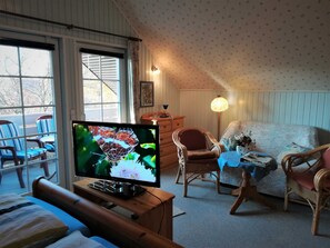 Ein gemütliches Zimmer mit Fernseher auf Holzständer, einem Bett mit floralem Überwurf, einem Sessel und einem Tisch mit Lampe.