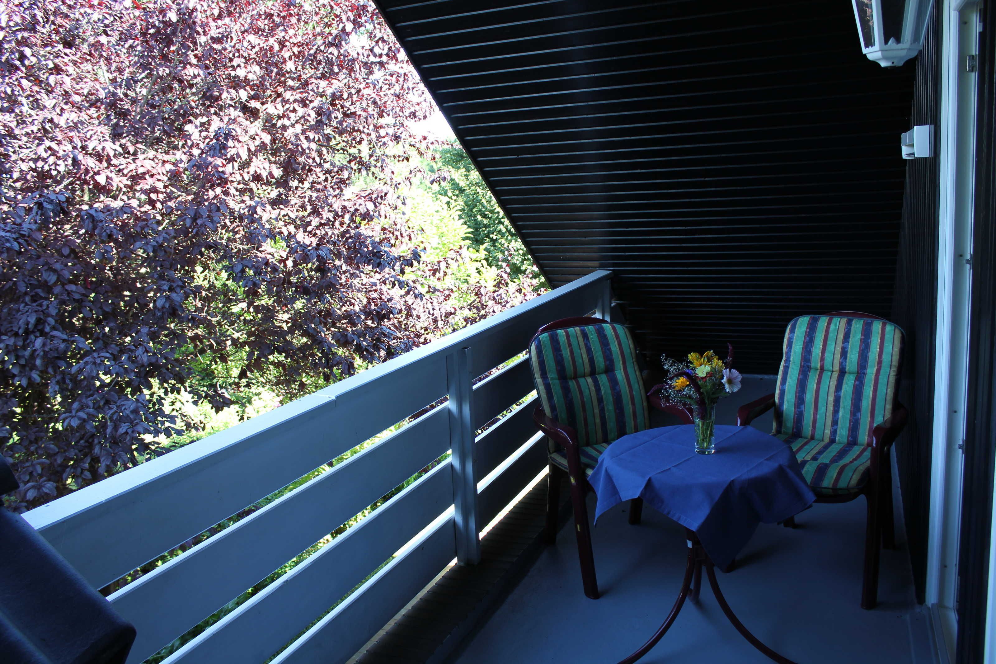 Ein Balkon mit Tisch, zwei gestreiften Stühlen, Geländer und Blick auf Grünpflanzen.