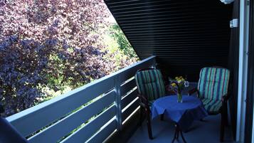 Ein Balkon mit Tisch, zwei gestreiften Stühlen, Geländer und Blick auf Grünpflanzen.