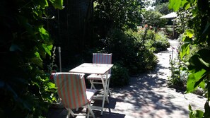 Eine Gartenpatio mit Tisch und zwei gestreiften Stühlen, umgeben von üppigem Grün und einem Ziegelweg.