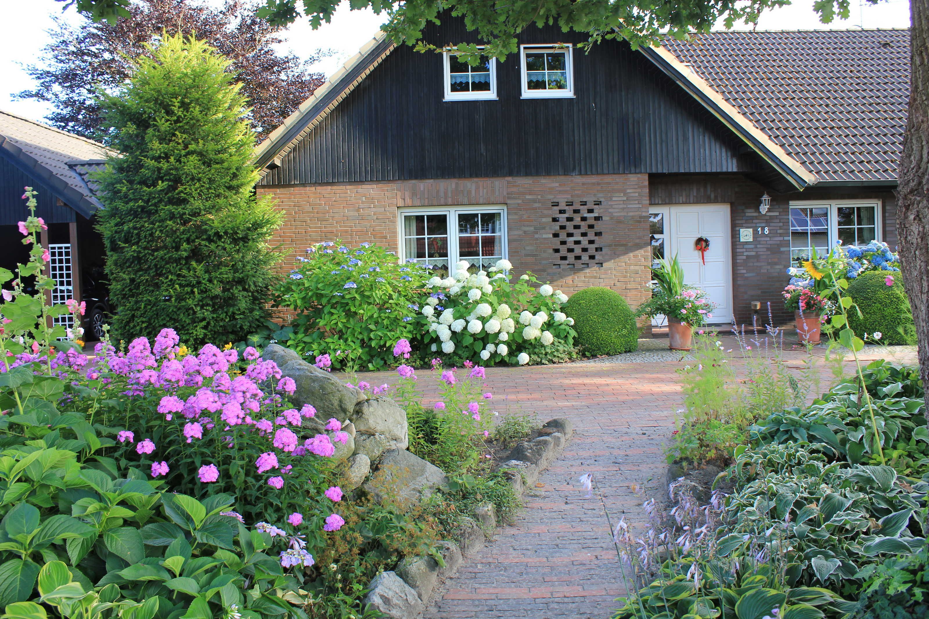 A house with a brick exterior, a dark roof, and a front door with a wreath. There are flowering plants and a pathway leading to the entrance.