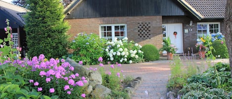 A house with a brick exterior, a dark roof, and a front door with a wreath. There are flowering plants and a pathway leading to the entrance.