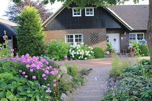 Ein Haus mit Backsteinfassade, dunklem Dach und einer Haustür mit einem Kranz. Vor den Blumen blühen und ein Weg führt zum Eingang.