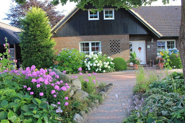 A house with a brick exterior, a dark roof, and a front door with a wreath. There are flowering plants and a pathway leading to the entrance.