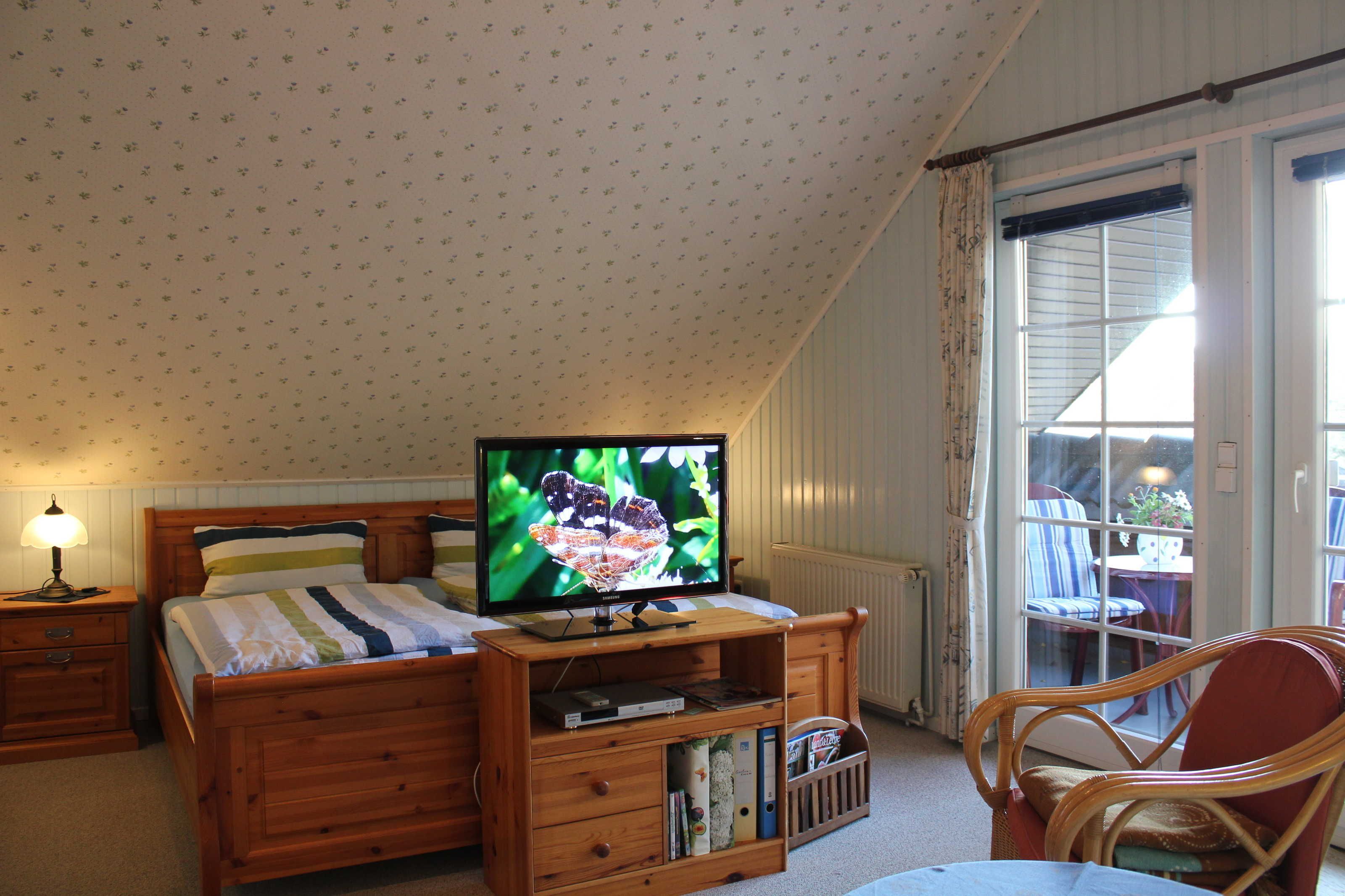 Ein Schlafzimmer mit einem Bett, einem Fernseher auf einem Holztisch, einem Nachttisch, einem Sessel und einem Fenster mit Vorhängen.