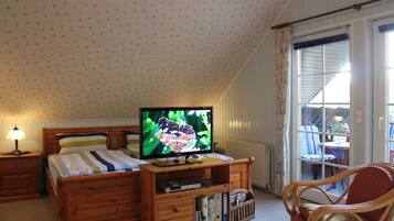 Ein Schlafzimmer mit einem Bett, einem Fernseher auf einem Holztisch, einem Nachttisch, einem Sessel und einem Fenster mit Vorhängen.