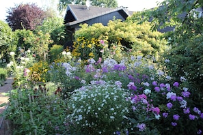 En trädgård med många olika sorters blommor och en byggnad i bakgrunden.