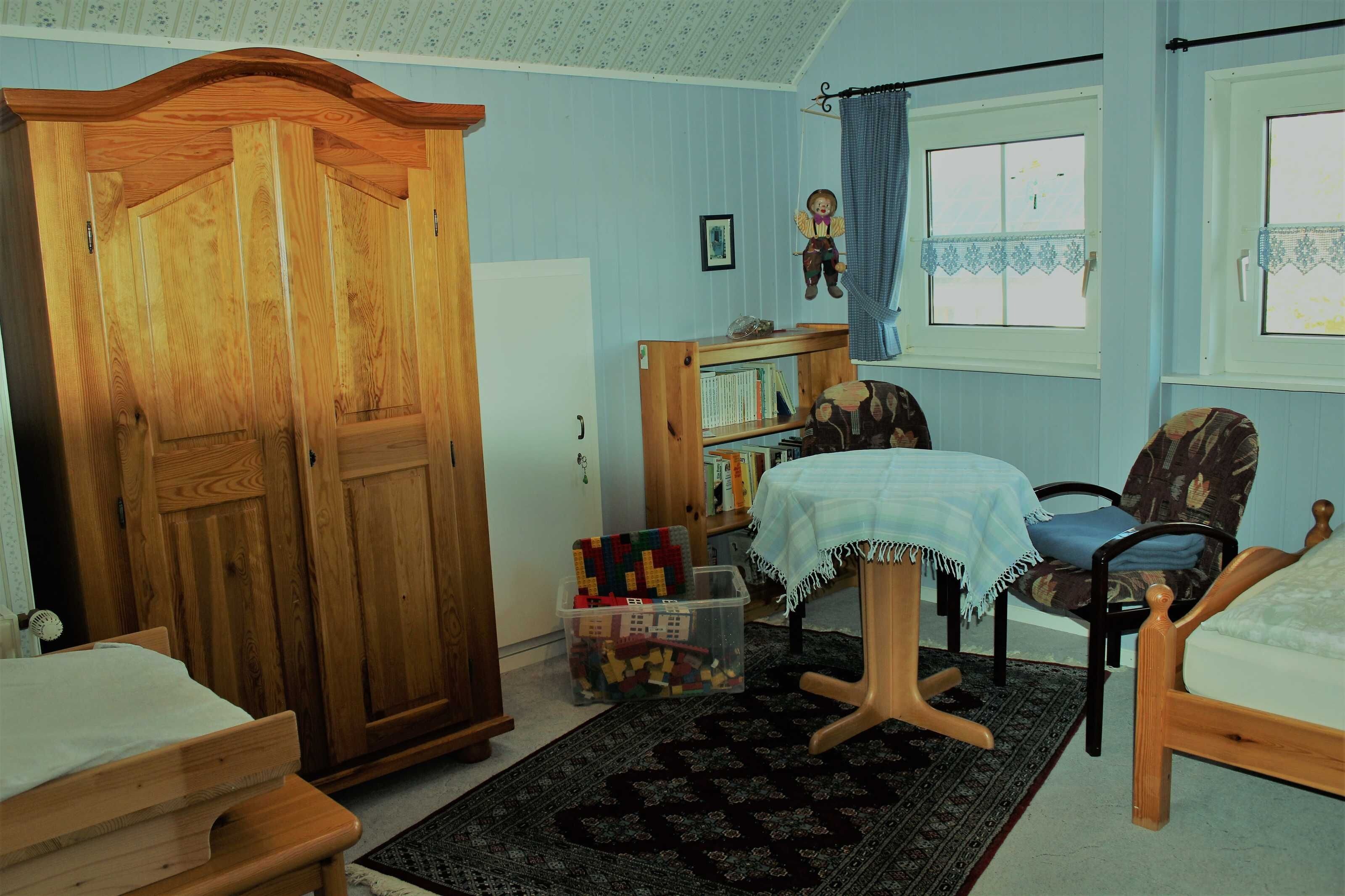 A room with a wooden wardrobe, a small table with a white tablecloth, a chair, a bed, and a bookshelf.
