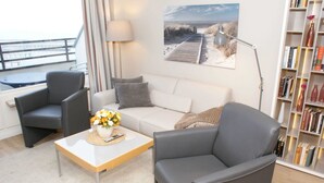 A modern living room with a sofa, two armchairs, a coffee table with a vase of flowers, a bookshelf, and a painting of a beach scene.