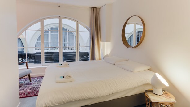 Hotelkamer met een groot bed, een bedlampje, een ronde spiegel en uitzicht op de stad door grote ramen.