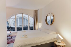 Ett hotellrum med en stor säng, en sänglampa, en rund spegel och utsikt över staden genom stora fönster.