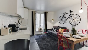 Een moderne keuken met witte kasten, een zwart aanrechtblad en een inbouwoven. Er is een eethoek met een houten tafel, rode stoelen en een grijze bank. Een fiets hangt aan de muur boven de eettafel.