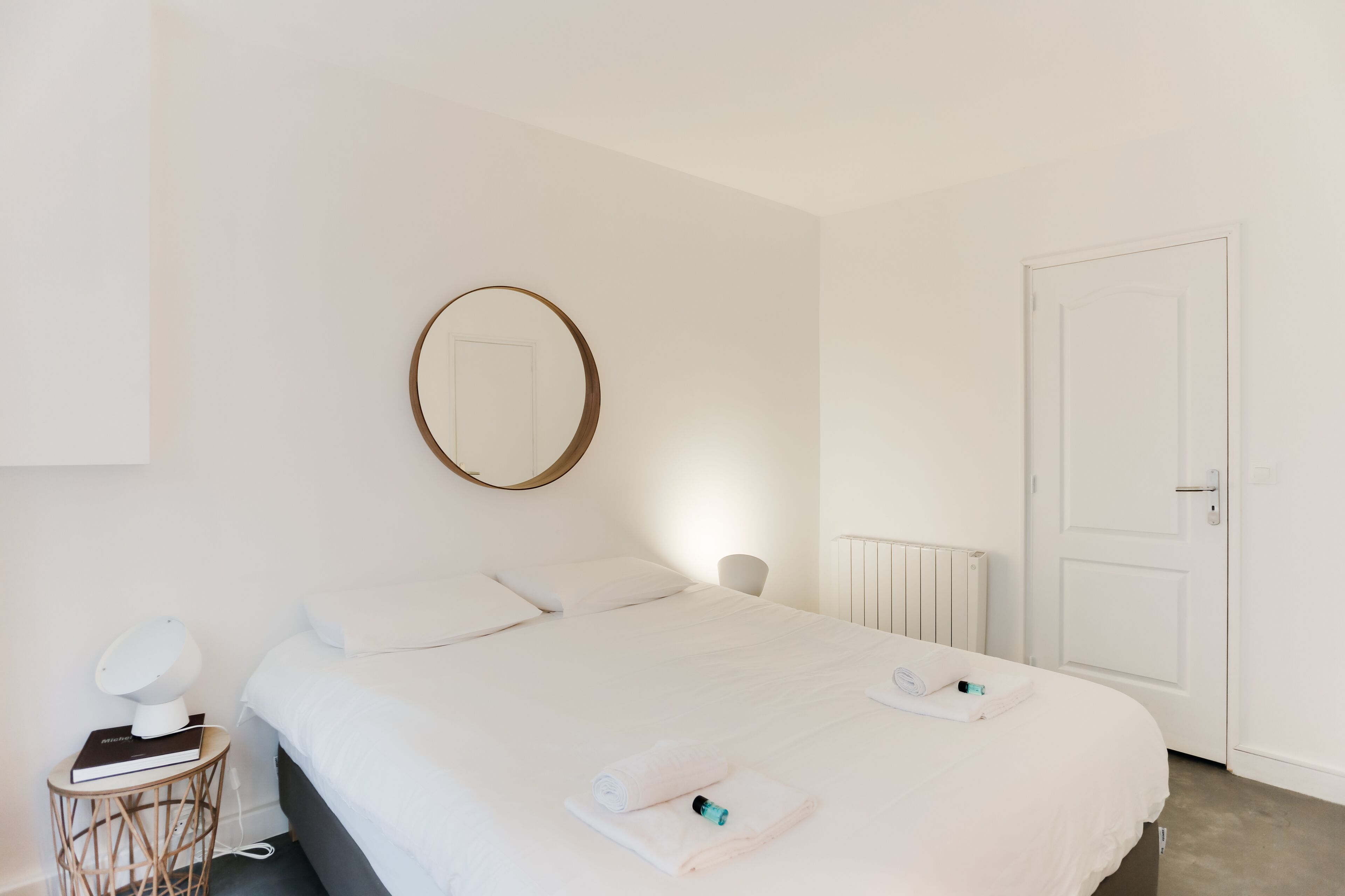 Ein schlicht eingerichtetes Hotelzimmer mit einem ordentlich bezogenen Bett, einem runden Spiegel an der Wand, einem Nachttisch mit Lampe und einer weißen Tür.