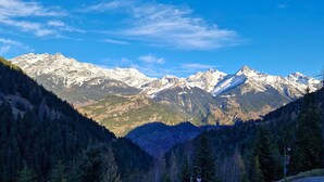 Paisaje montañoso con picos nevados, un bosque denso y una carretera pavimentada con una cerca de madera.