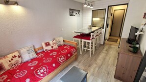 A cozy living area with a sofa, a dining table, and chairs. There is a small table in the foreground and a television on a stand to the right. - 2-Room + Cabin Apartment for 4-6 in Heart of Station - WiFi & Balcony (Valfréjus)
